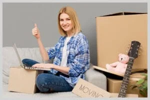 Smiling woman with laptop giving thumbs up while surrounded by moving boxes